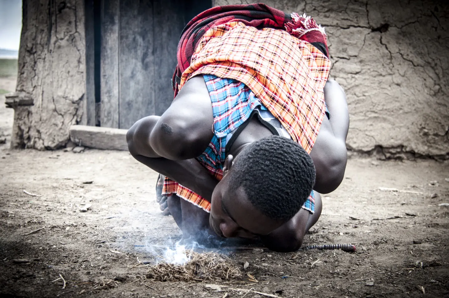 A Maasai warrior in a red shúkà kneels to light a fire by hand