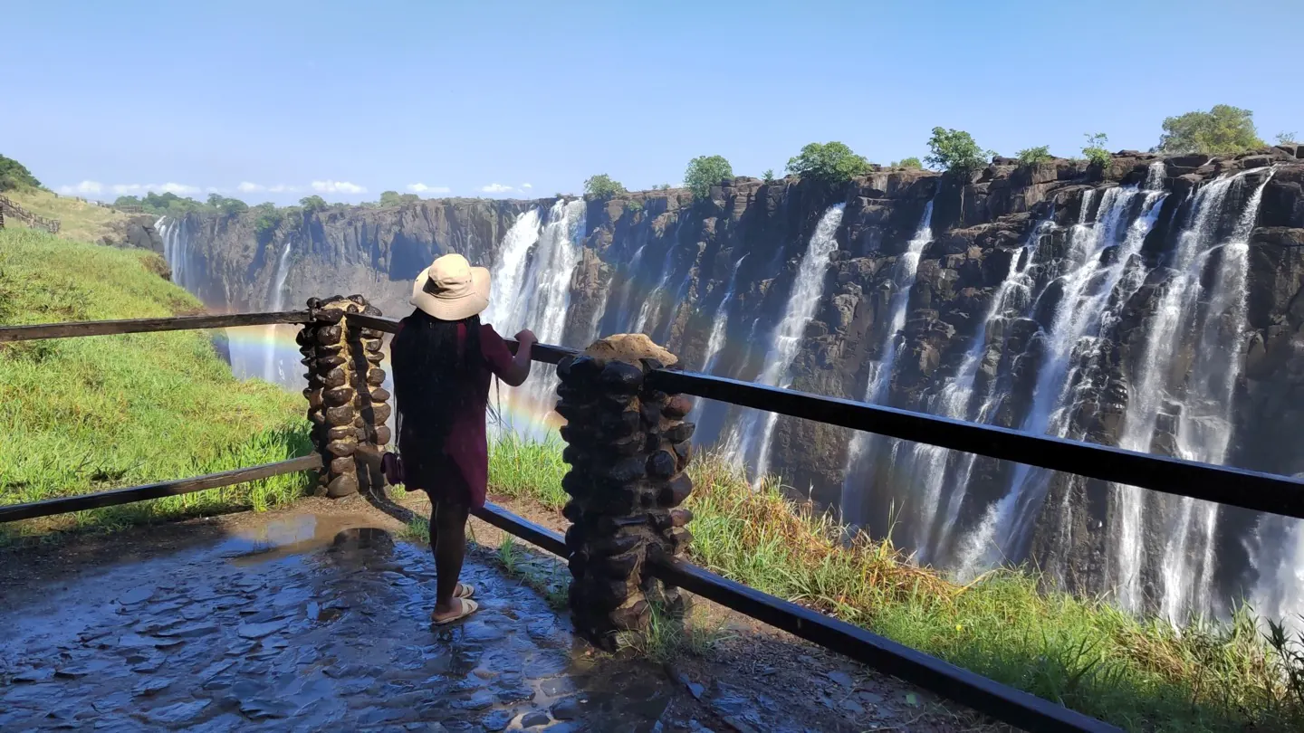 A traveller in a sun hat looks out over Victoria Falls, with a rainbow in the spray